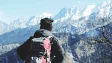 photography of man in black hooded jacket and red backpack facing snow covered mountain